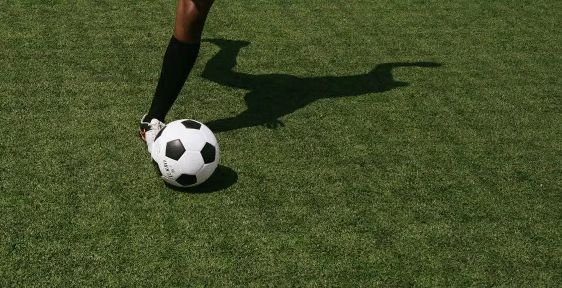 Pernas de uma mulher chutando uma bola em um campo de futebol.